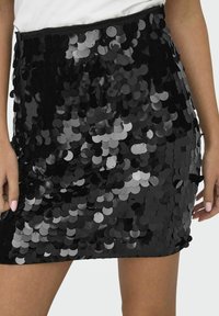 Mini falda de lentejuelas negras con lentejuelas reflectantes circulares de diferentes tamaños, creando una superficie texturizada y brillante sobre una base de tela sólida.