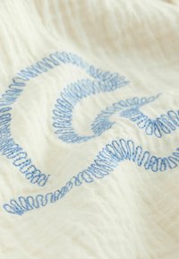 Weißer strukturierter Stoff mit lockerem blauem Schlaufenstickmuster, das ein wellenförmiges, fließendes Muster über die Oberfläche bildet.