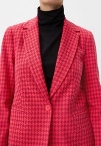 Czerwony żakiet w wzór houndstooth z pojedynczym zapięciem na guzik, zwężonym kołnierzem i teksturowanym materiałem, założony na czarny golf.