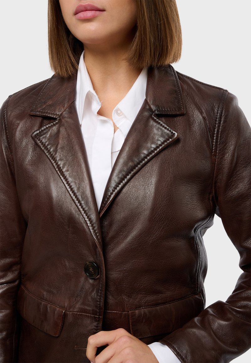 Giacca in pelle marrone con rever affilati, chiusura a un bottone e finitura testurizzata. Indossata sopra una camicia bianca.