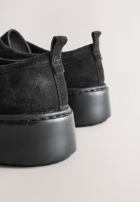 Sorte mokkasinlignende sko med tekstureret finish, med en robust sort gummisål og små bageste hælkiler for nem påtagning.