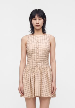 Jeune femme aux cheveux noirs portant une robe sans manches beige à carreaux avec des boutons devant, debout sur un fond uni.