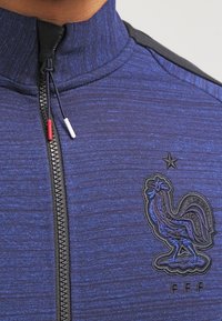 Giacca sportiva blu con zip, dotata di un colletto alto, realizzata in tessuto testurizzato, con zip nera e un logo di gallo ricamato con stelle.