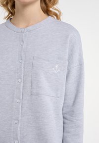 Cardigan grigio chiaro realizzato in tessuto morbido, con scollatura rotonda, bottoni frontali e una tasca sul petto con un dettaglio di ancora ricamato.