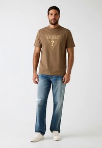 T-shirt marron à manches courtes avec un imprimé triangulaire doré et un logo, associé à un jean bleu clair déchiré et des baskets blanches.