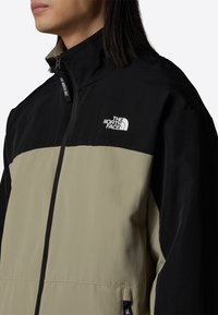 Personne portant une veste zippée beige et noire avec le logo The North Face sur la poitrine et le curseur de la fermeture éclair.