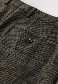 Nærbillede af uldbukser med ternet mønster i grønt og brunt samt en baglomme sikret med en mørk knap.