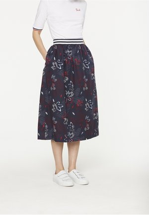 Femme portant une chemise blanche à manches courtes avec le texte « think! », une jupe midi bleu marine à motifs floraux avec des poches, et des baskets blanches à scratch, debout les mains dans les poches.