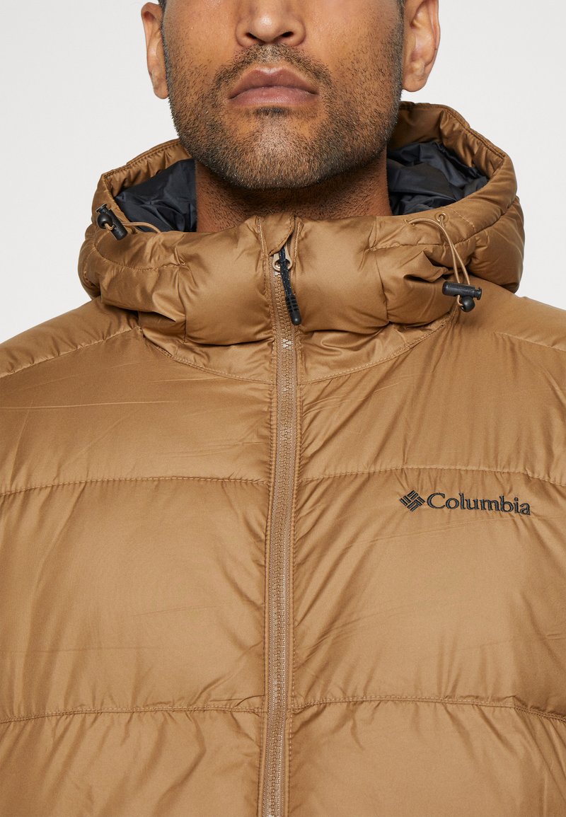 Jasnobrązowa puchowa kurtka z wysokim kołnierzem, zamkiem błyskawicznym z przodu i kapturem ze sznurkiem. Posiada logo Columbii na klatce piersiowej. Gładka tekstura.