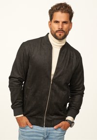 Schwarze Wildleder-Bomberjacke mit rundem Kragen, Reißverschluss vorne und gerippten Bündchen. Getragen über einem cremefarbenen Rollkragenpullover und kombiniert mit blauen Jeans.