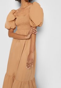 Brun klänning med puffärmar, smockad midja och lager på lager kjol. Tillverkad av lätt tyg; mjuk textur och flödande silhuett.