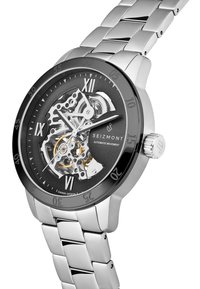 Automatisk klokke med sølvfarget rustfritt stålarmbånd og svart urskive. Har synlige gir, sølvfargede detaljer og romerske tall.