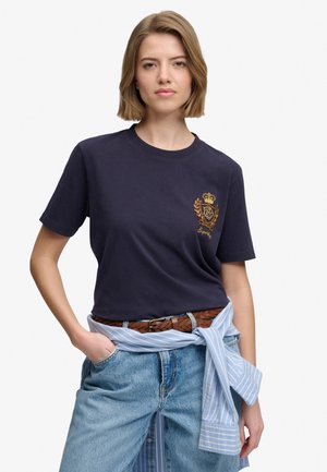 Frau trägt ein dunkelblaues T-Shirt mit besticktem Wappen, hellblaue Jeans und ein blau gestreiftes Hemd, das um ihre Taille gebunden ist, steht mit einer Hand in der Tasche.