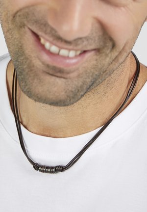 Braune Lederkette mit mehreren dunklen Perlen, die an einer schwarzen Schnur hängt. Wird über einem schlichten weißen T-Shirt getragen, Nahaufnahme am Ausschnitt.