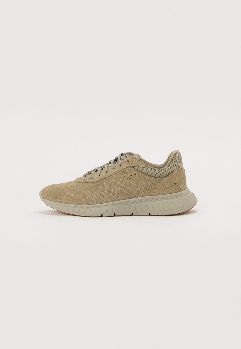 Beige zamšas sneakers ar acs akcentiem, noapaļota purngala, plakaniem šņorēm un kontrastējošu gumijas zoli. Izceļas ar tekstūru un ventilācijas detaļām.