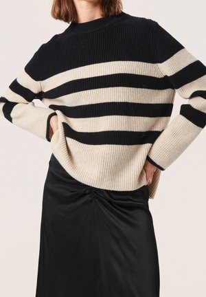 Personne portant un pull à rayures horizontales noir et beige associé à une jupe midi noire froncée, les mains posées sur les hanches.