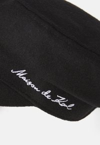 Béret en laine noir avec une forme arrondie, ornée du texte brodé en blanc "Maison de Zal" le long du bord. Texture lisse avec des accents minimaux.