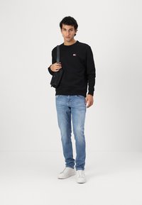 Black sweatshirt with a small logo, blue jeans, and white sneakers. The model holds a black crossbody bag. Simple, casual outfit.