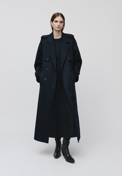 Granatowy płaszcz trench z podwójnym zapięciem, dużymi klapami i bocznymi kieszeniami; połączony z czarną sukienką i czarnymi botkami za kostkę.