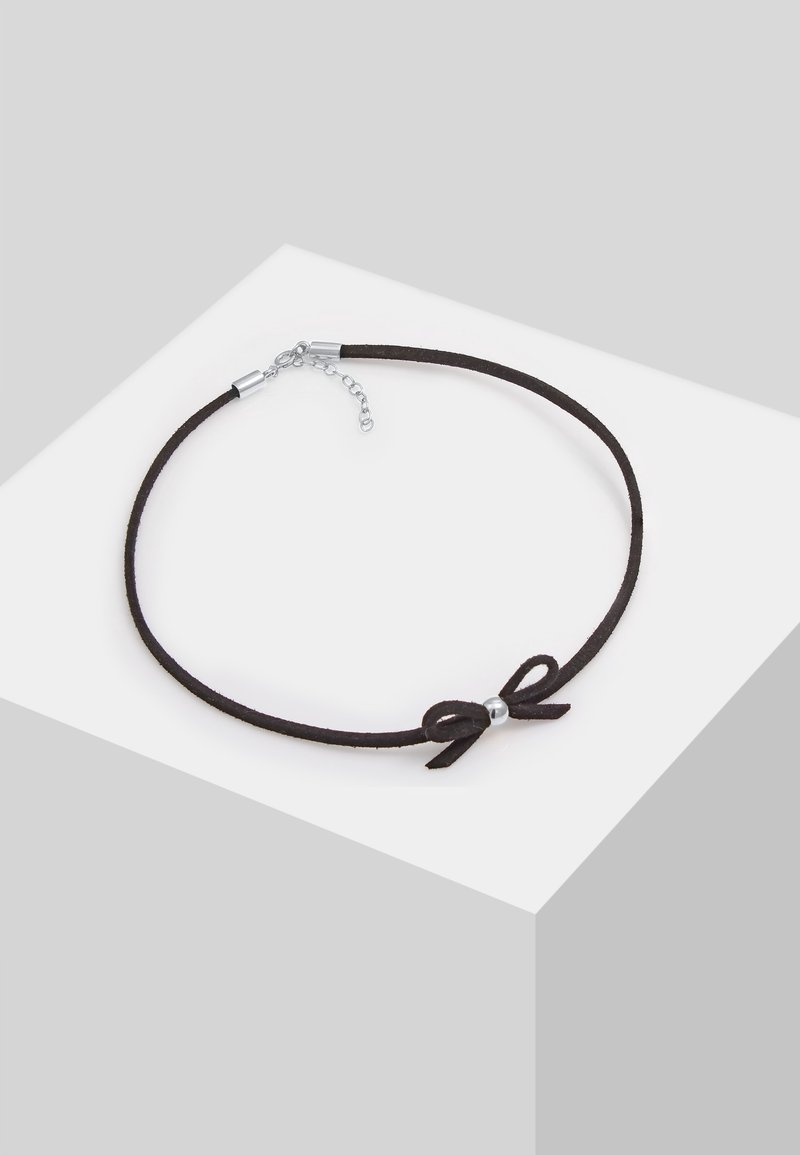 Collier ras-du-cou en daim noir avec un petit nœud et une perle argentée au centre, présenté sur une surface blanche et grise.