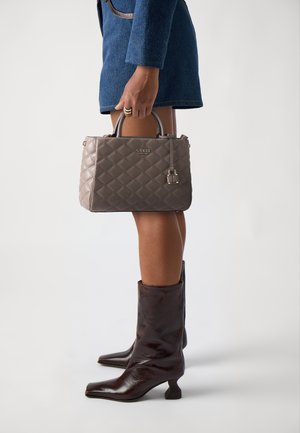 Bolso acolchado gris con asas superiores, que presenta un detalle de cierre y un logo en relieve. El bolso se sostiene junto a unas botas de tacón alto marrones.