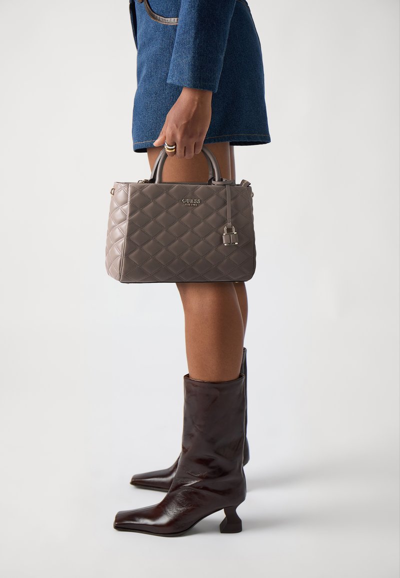 Bolso acolchado gris con asas superiores, que presenta un detalle de cierre y un logo en relieve. El bolso se sostiene junto a unas botas de tacón alto marrones.