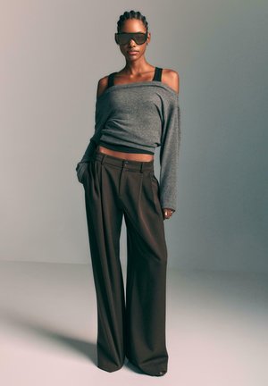 Grå off-shoulder sweater med lange ærmer, parret med løse, højhalsede brune bukser. Model iført store sorte solbriller.