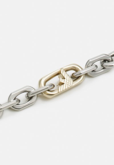 Corrente de metal com elos prateados alternados e um destaque em ouro. O elo dourado tem detalhes em relevo e um padrão triangular.