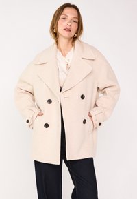 Cappotto doppio petto color beige chiaro con ampi rever, bottoni neri, una vestibilità rilassata e tasche laterali. Tessuto morbido e strutturato.