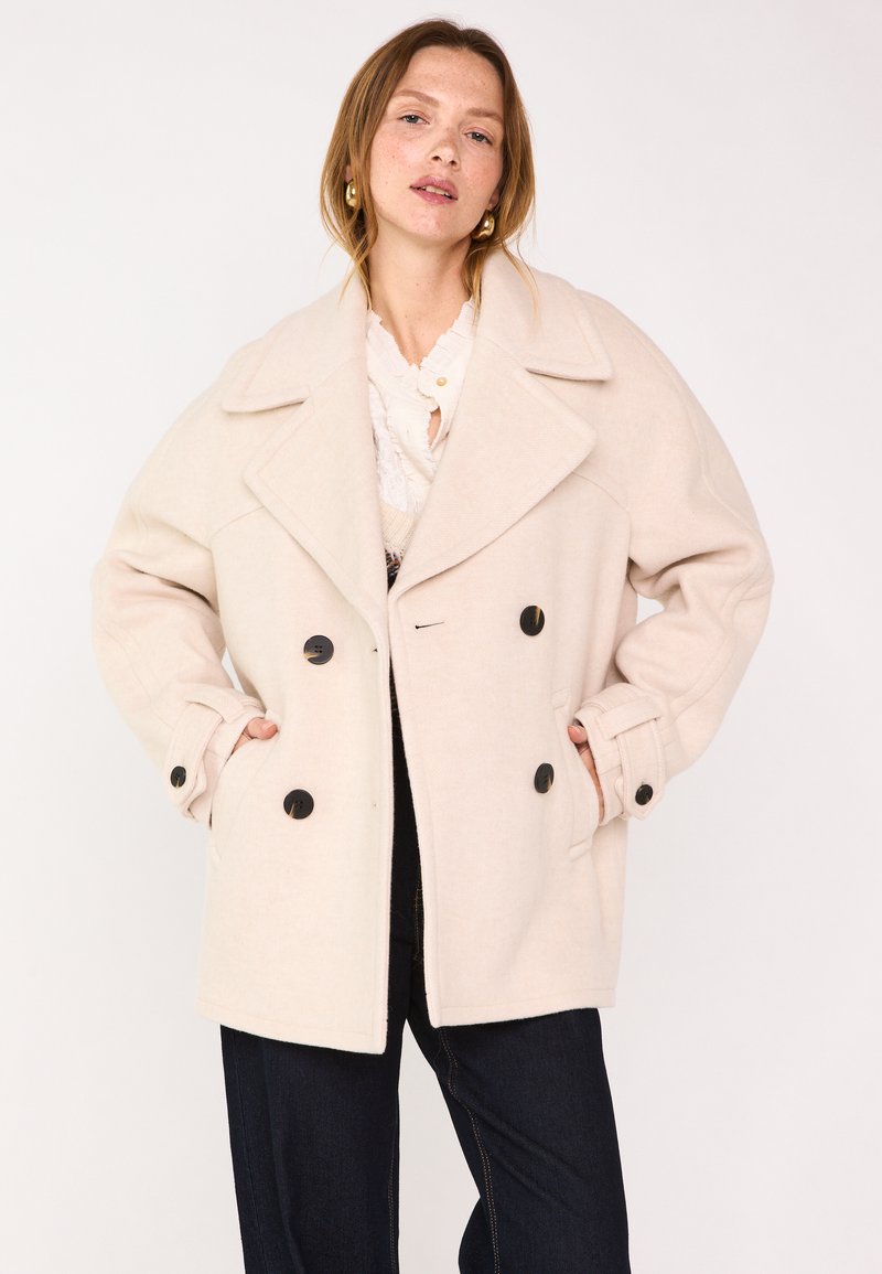 Cappotto doppio petto color beige chiaro con ampi rever, bottoni neri, una vestibilità rilassata e tasche laterali. Tessuto morbido e strutturato.