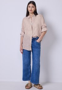 Giovane donna in piedi con una mano in tasca, indossa una camicia beige a righe con bottoni, jeans blu a gamba larga e sandali beige.