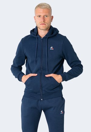 Sudadera con cremallera en color marino, con un bolsillo tipo canguro, capucha ajustable con cordón y parche con logo en el pecho. Tejido suave de peso medio.
