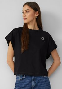 Musta puuvillainen lyhythihainen crop-top, jossa on pyöreä kaula-aukko, ja vasemmassa rinnassa valkoinen graafinen kuvio.