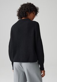 Maglione nero a coste con una vestibilità ampia, scollo rotondo e maniche lunghe, abbinato a pantaloni grigi chiari. Tessuto con texture visibile.