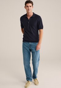 Jauns vīrietis stāv, ģērbies tumši zila īsām piedurknēm polo kreklā, zilās džinsās un bēšos zamšādas apavos, pret vienkāršu, gaišu fonu.