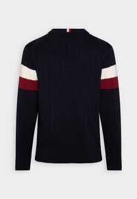 Pull côtelé bleu marine à manches longues, avec des rayures horizontales blanches et rouges sur le haut des bras et une petite rayure rouge, blanche et bleue au niveau du col.