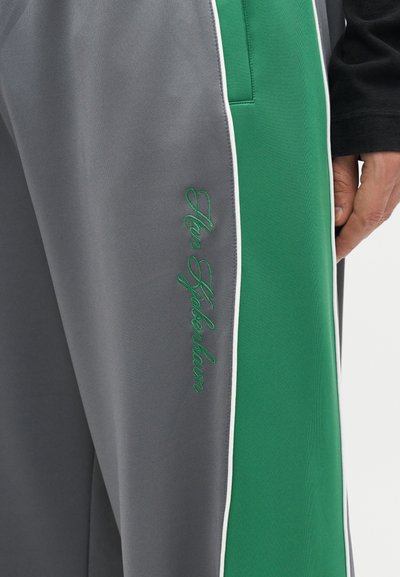 Γκρι και πράσινο παντελόνι με λευκές ραφές και πράσινη καλλιγραφική κέντημα που γράφει «Hen Hickenbaux» κοντά στην τσέπη, φορεμένο από ένα άτομο με ορατό χέρι.