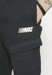 Černé tepláky Nike s postranní kapsou, jednolitým vzorem a bílým logem Nike se třemi diagonálními pruhy nad ním.