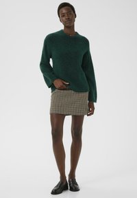Vrouw in een donkergroene fuzzy trui, een geruite minirok en zwarte instappers, staand tegen een eenvoudige lichtgrijze achtergrond.