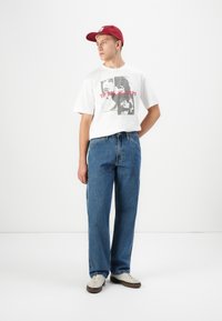 Ung mand iført en rød kasket, en hvid t-shirt med Rolling Stones grafik, blå jeans og beige sneakers, stående mod en ensfarvet hvid baggrund.