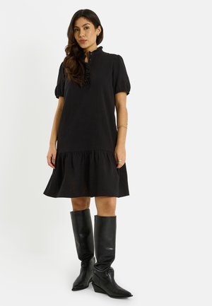 Femme aux cheveux longs et foncés portant une robe noire à manches courtes avec des détails à volants et des bottes en cuir noires montant jusqu'aux genoux, debout devant un fond blanc.