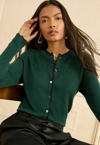 Tmavozelený kardigán so zúbkovaným výstrihom, textúrovanou prednou časťou a striebornými gombíkmi. Čierne akcenty na golieri a manžetách. Hladký pletený materiál.