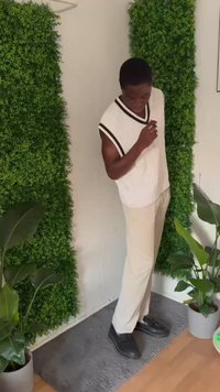 White sleeveless sweater with black stripes, beige pants, and black shoes. Surrounded by greenery and indoor plants against a textured backdrop.