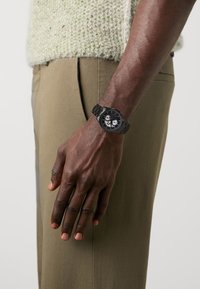 Orologio analogico nero con cinturino nero testurizzato, caratterizzato da un quadrante rotondo con sotto-quadranti e marcatori bianchi a contrasto, mostrato su una mano.