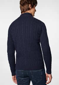 Granatowy sweter w splot żakardowy z wysokim kołnierzem, ściągaczami przy mankietach i u dołu. Charakteryzuje się teksturowanym wzorem dla dodatkowej głębi. Noszony z ciemnym dżinsem.