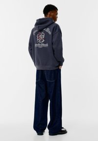 Hanorac de un gri închis cu grafică pe spate, având text și un design floral. Asortat cu pantaloni din denim întunecat, cu croială lejeră. Pantofi negri cu talpă plată.