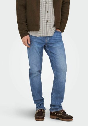 Man draagt lichtblauwe rechte jeans, een bruine jas, geruit overhemd en donkerbruine leren bootschoenen, staande met één hand in de zak.