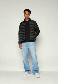 Giacca nera imbottita con cerniera frontale e colletto alto, abbinata a una maglietta nera e jeans azzurri chiari, indossata con sneakers bianche.