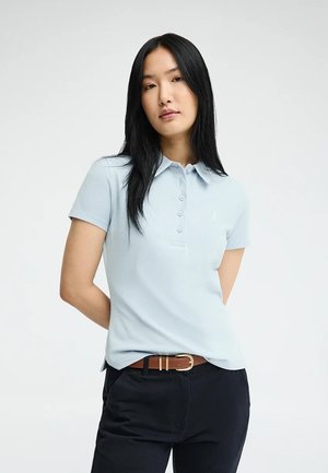 Mujer con cabello largo y negro, vistiendo una camisa polo de manga corta azul claro y pantalones negros con un cinturón marrón, de pie frente a un fondo liso.