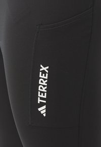 Leggings de sport noirs avec une texture lisse, dotés d'une poche latérale et d'un logo "TERREX" blanc sur la cuisse.